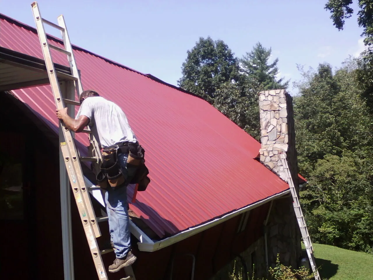 Roofer on a ladder beside a red metal roof and stone chimney — Soffit & Fascia Repair by Roofing Brothers in Otis Orchards